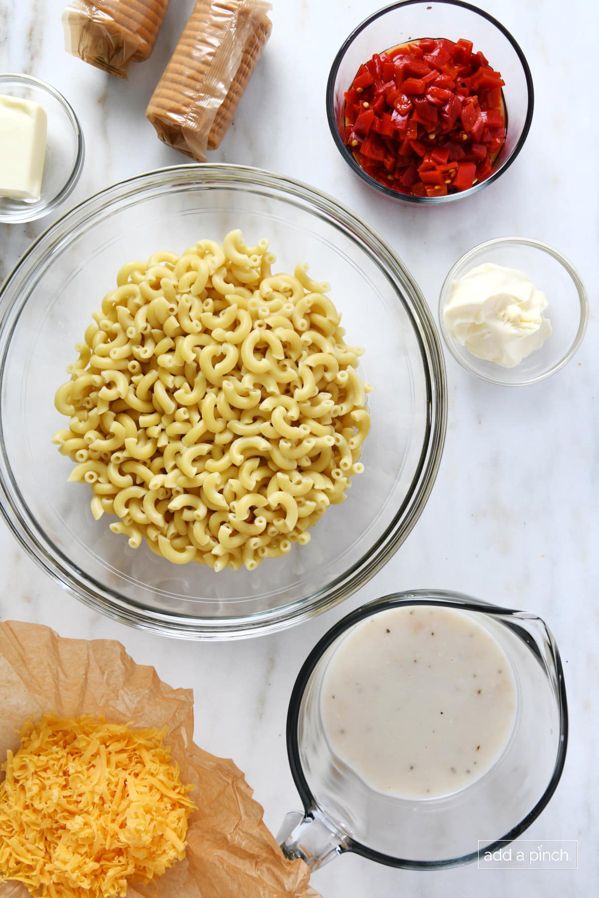 Glass bowls with ingredients for mac and cheese - butter, pimentos, mayo,  cream of chicken soup, elbow noodles, along with sleeves of crackers and mound of shredded cheese.