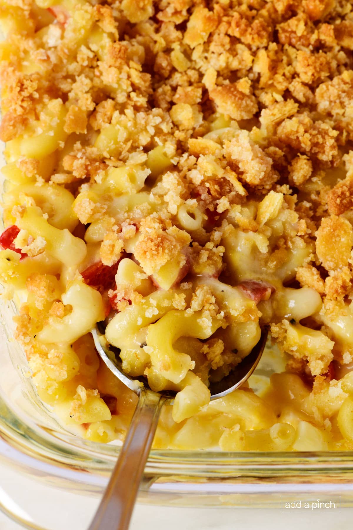 Closeup of macaroni and cheese in glass dish with spoon. Pimentos and cracker topping are visible with macaroni pasta and cheese.