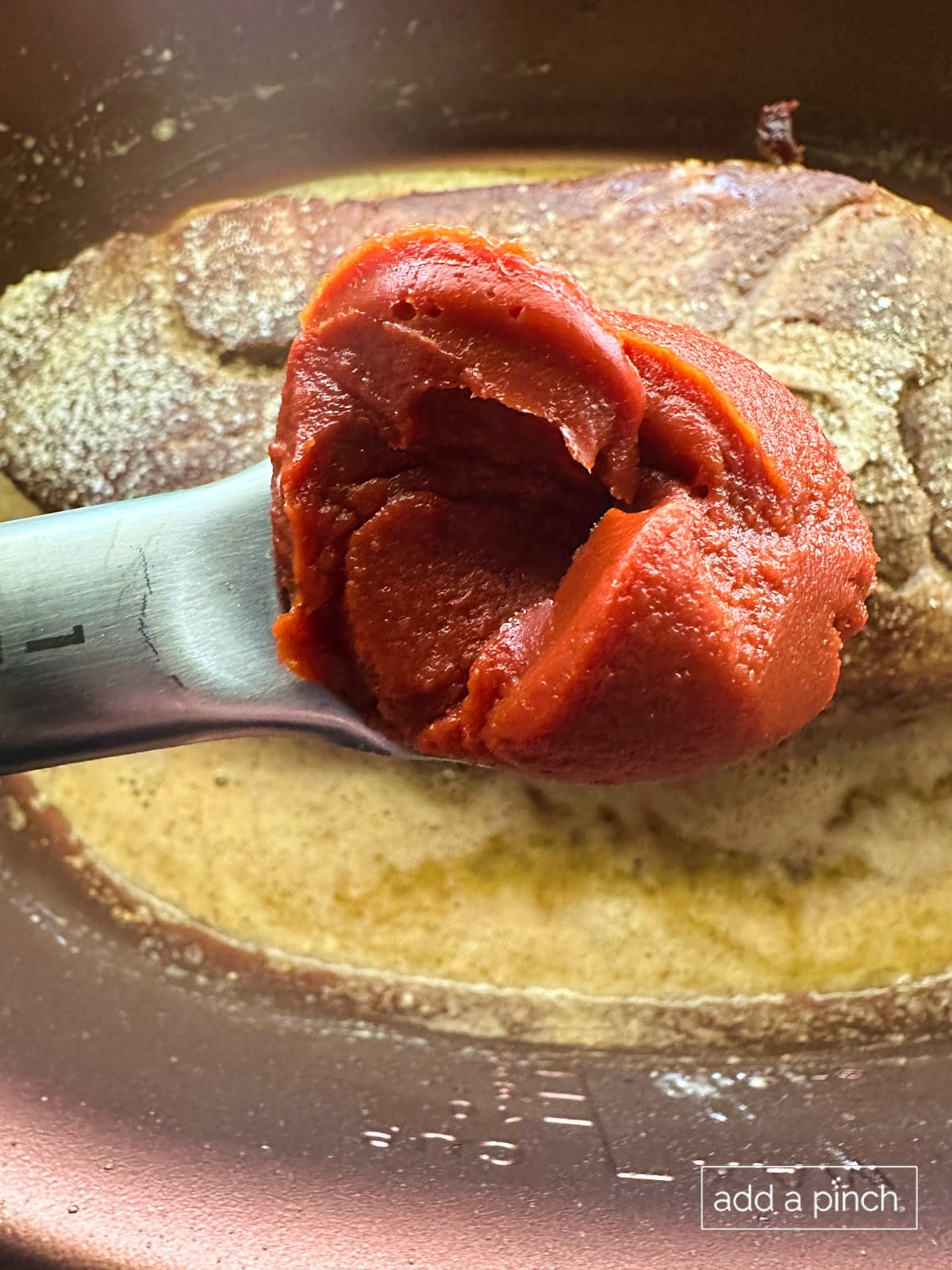 Tomato paste on measuring spoon being added to slow cooker.