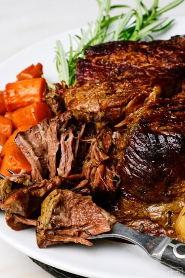 Pot roast on a white platter with vegetables and rosemary.