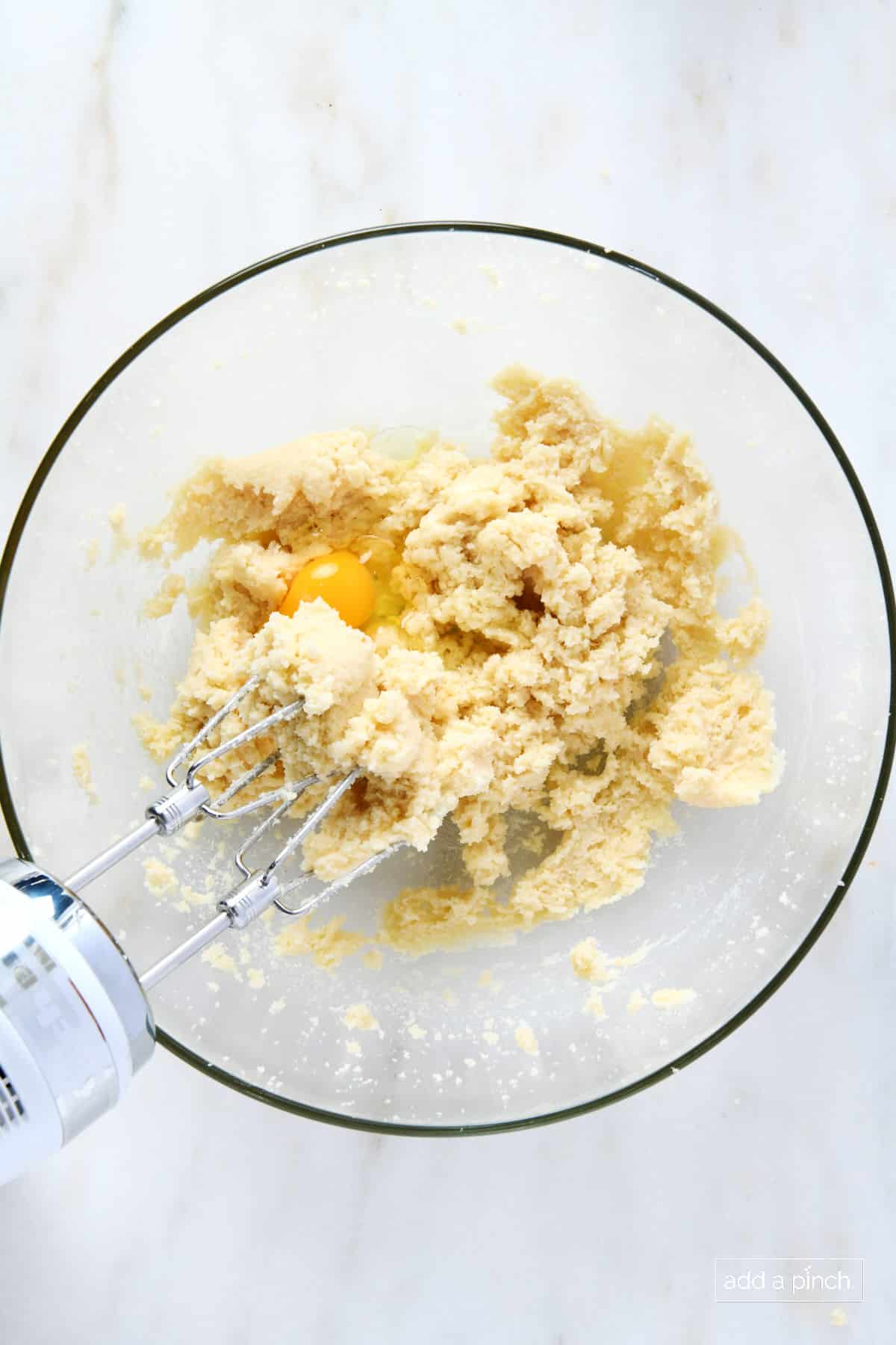Egg added to sugar cookie dough in a glass bowl with hand mixer.