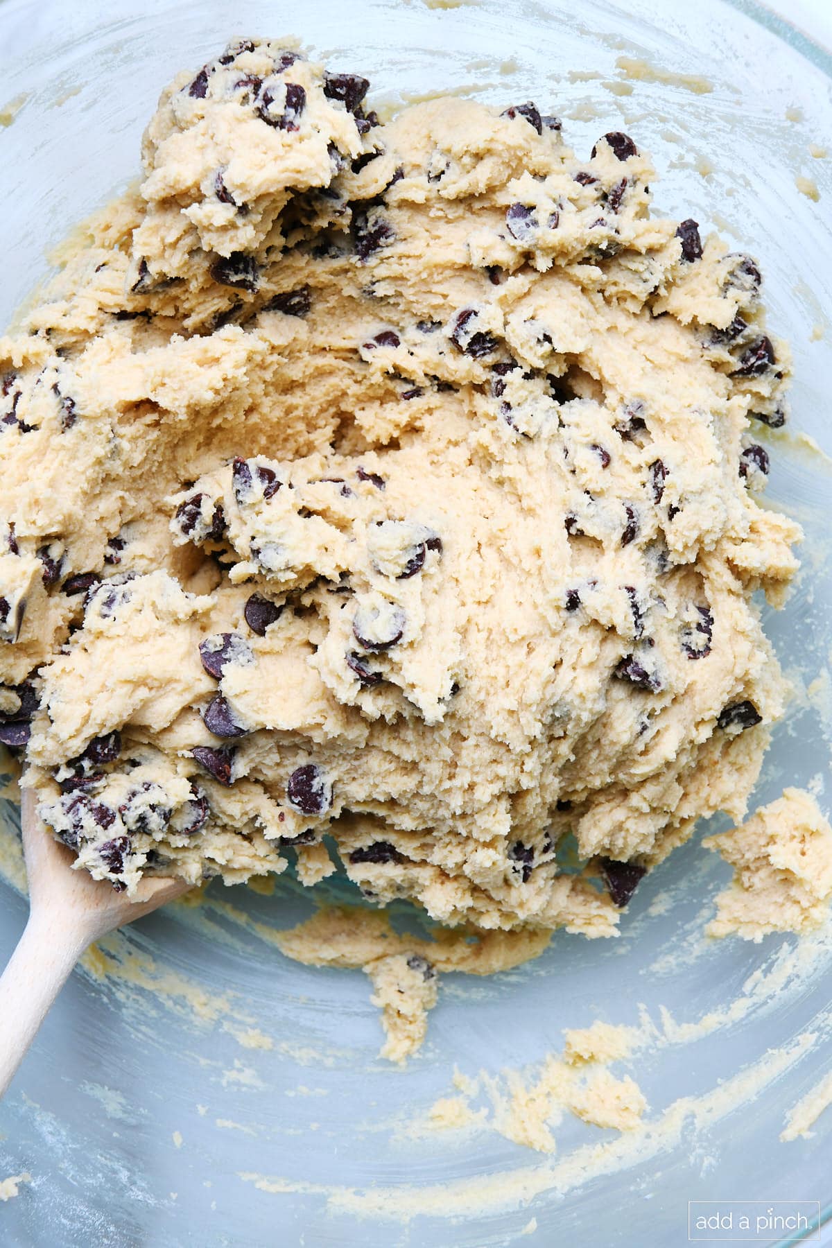 Chocolate chips blended into cookie cake dough in glass bowl with wooded spoon.