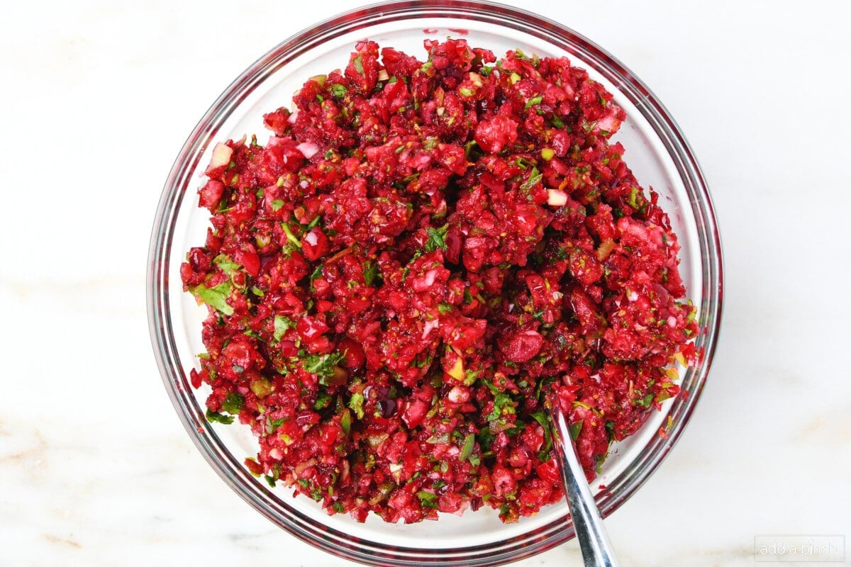 Cranberry Salsa and a spoon in the food processor. Cilantro, jalapenos, coarsely chopped cranberries, and orange zest are visible in the salsa mixture.