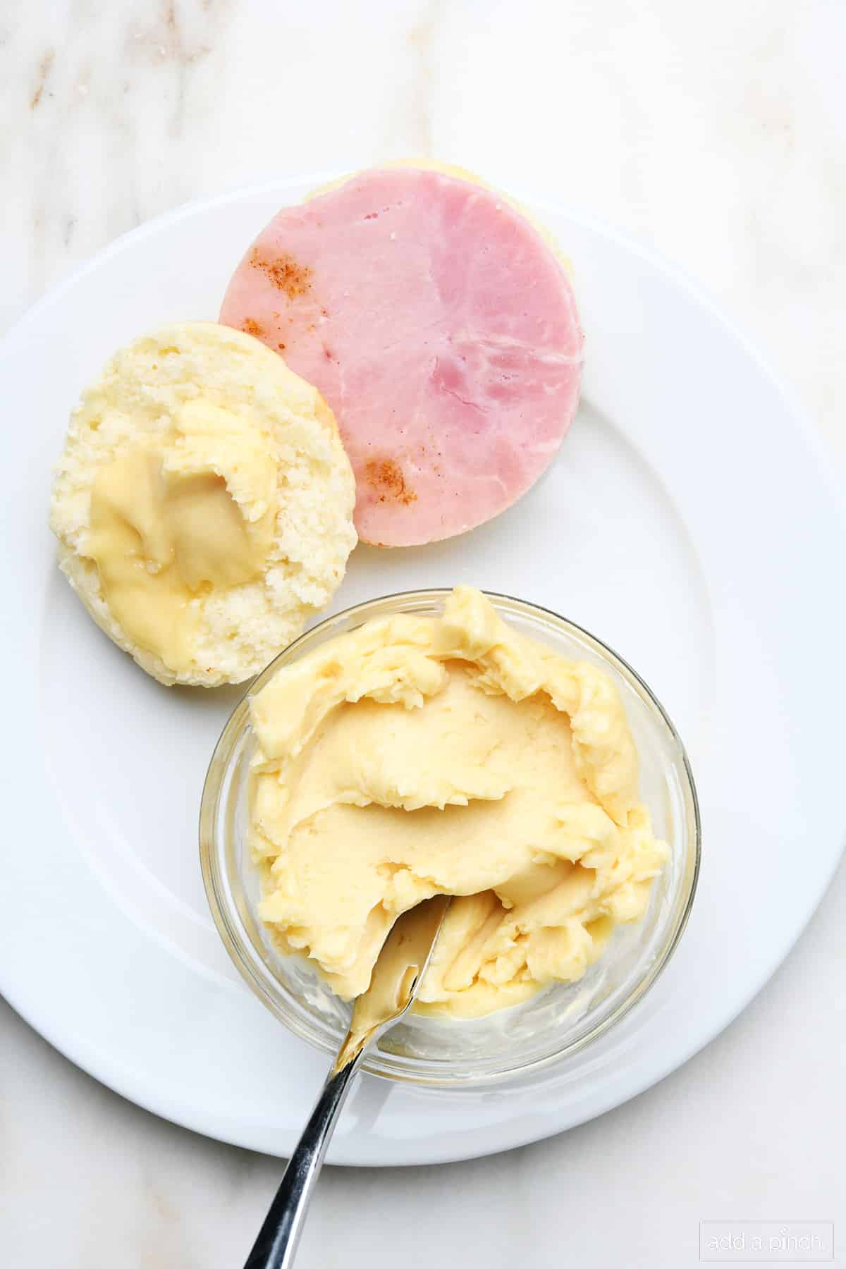 Honey butter in a glass bowl with butter knife and spread onto the top of a biscuit with ham on the side of the plate.