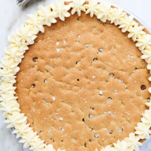 Vanilla buttercream frosting has been piped in star flower shapes around the edge of this chocolate chip cookie cake. Piping bag full of frosting is next to cake, all on marble countertop.