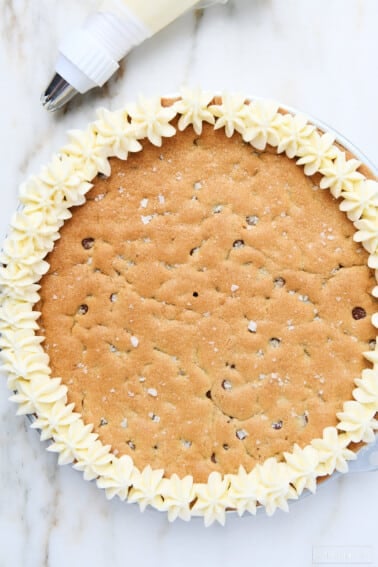 Vanilla buttercream frosting has been piped in star flower shapes around the edge of this chocolate chip cookie cake. Piping bag full of frosting is next to cake, all on marble countertop.