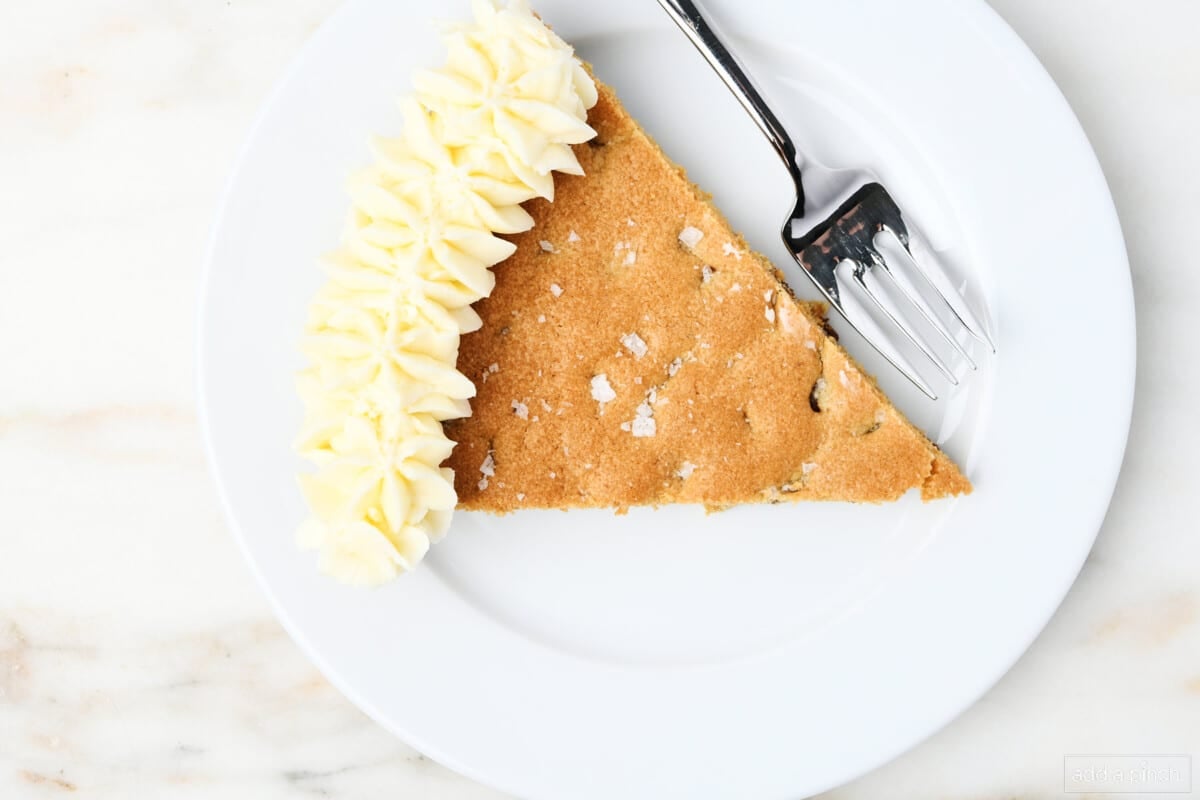 Overhead picture of slice of cookie cake with vanilla buttercream frosting and sprinkled with flake salt on a white dessert plate with fork.