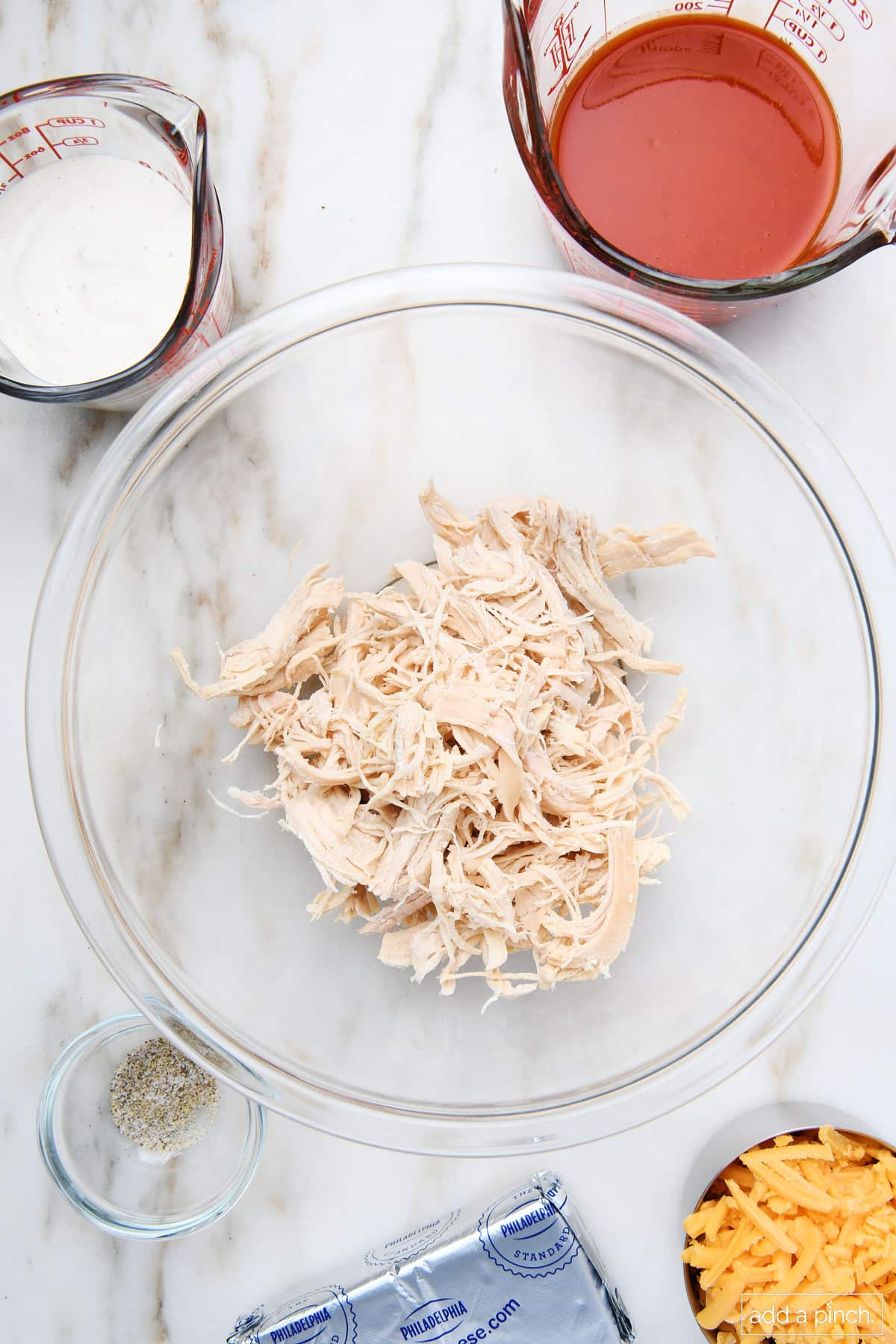 Ingredients used to make buffalo chicken dip on a marble surface. Shredded cooked chicken, ranch dressing, buffalo sauce, Stone House Seasoning, cream cheese, and shredded cheese. 