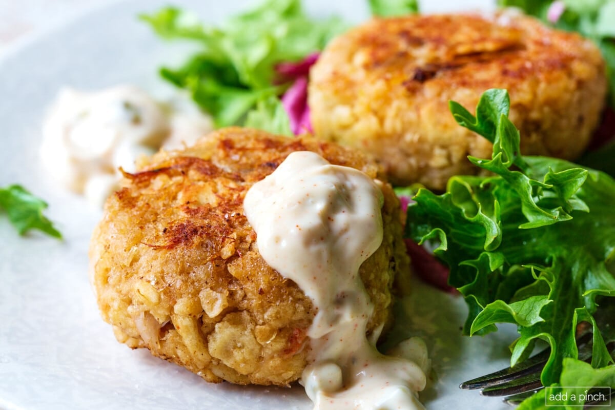 Two golden crab cakes on top of lettuces and drizzled with sauce. 