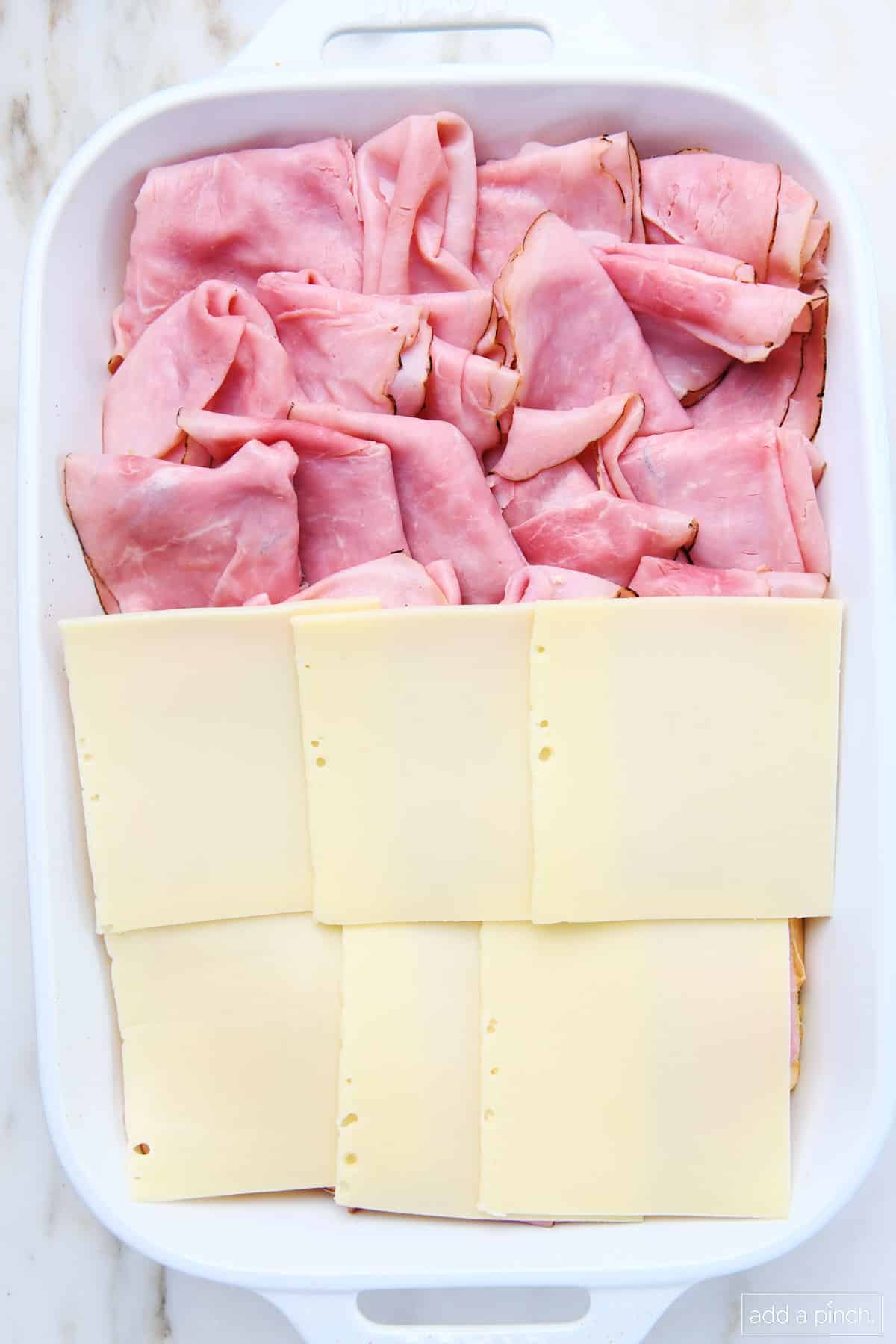 Swiss cheese being layered onto slices of ham and rolls in a 9x13 baking dish.