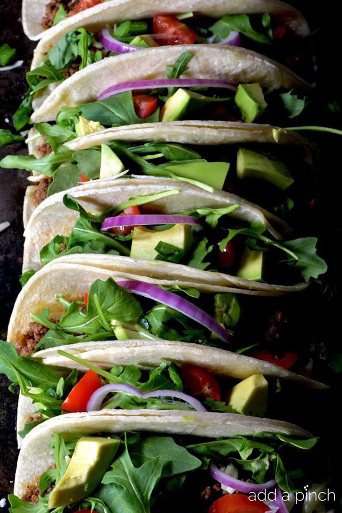 Row of soft beef soft shell tacos made with flour tortillas and filled with seasoned beef, lettuce, tomatoes, avocados and onions.