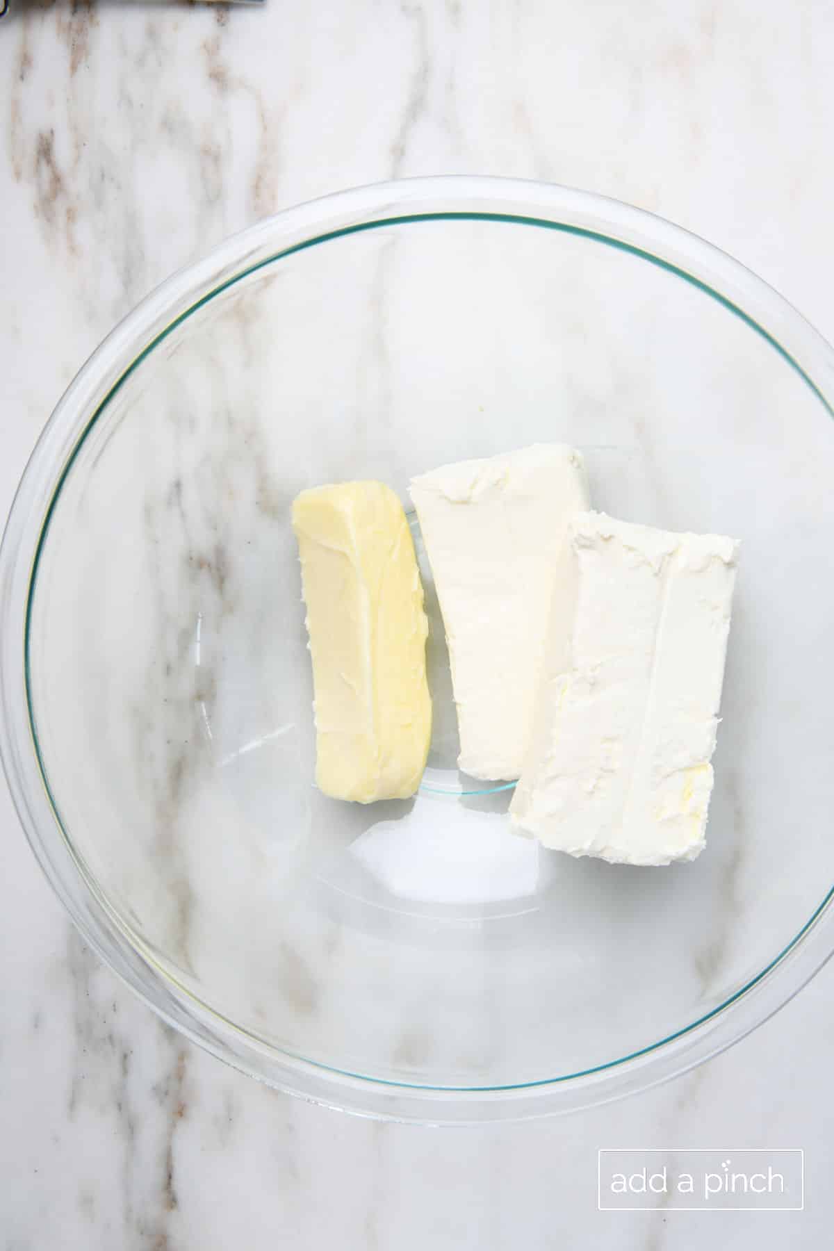 Two softened blocks of cream cheese and softened butter in a glass mixing bowl on marble countertop.