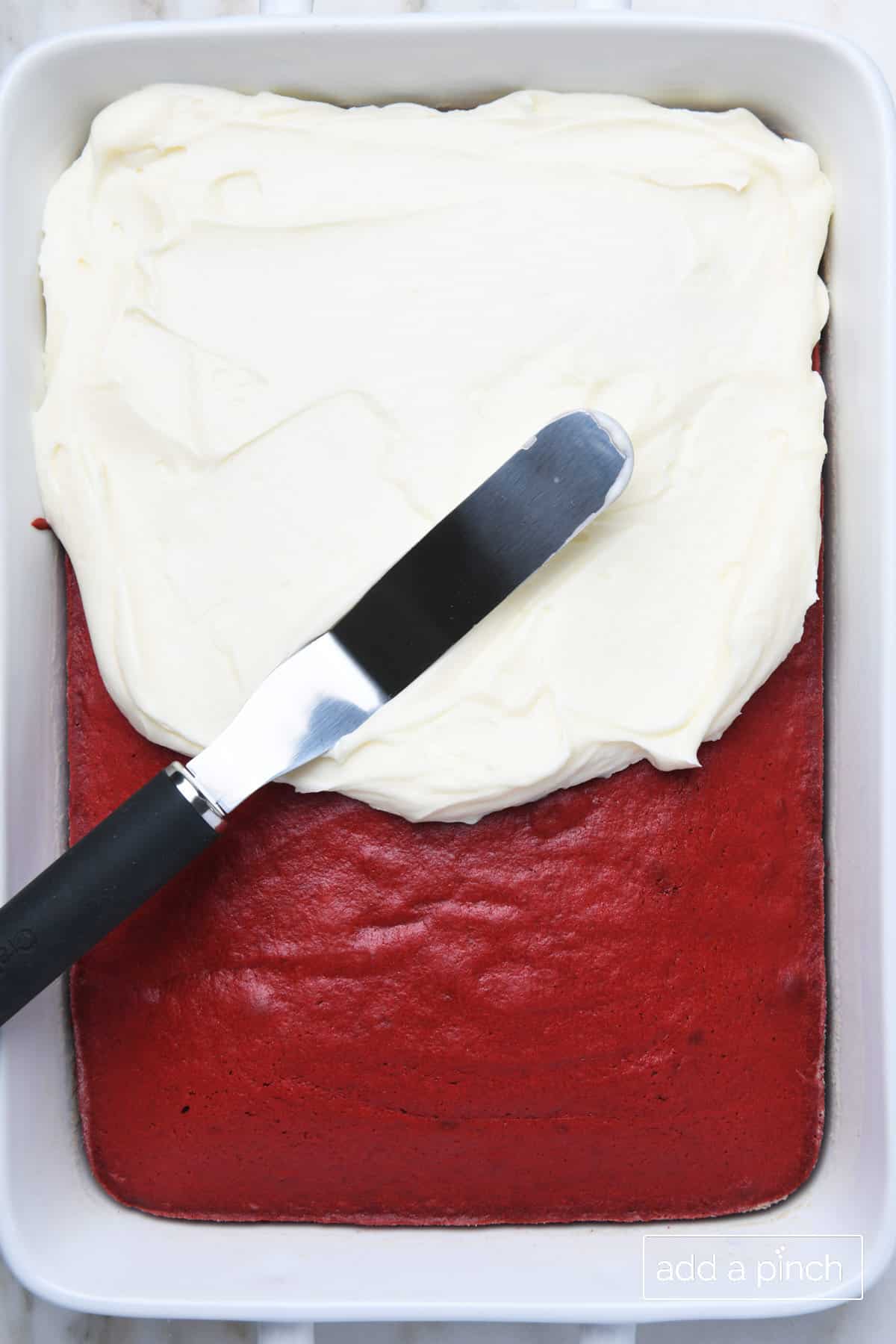 Cream cheese frosting being spread with an offset spatula onto a Red Velvet sheet cake.