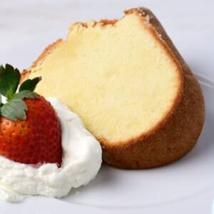 Slice of cream cheese pound cake with dollop of homemade whipped cream and red strawberry on a white dessert plate.