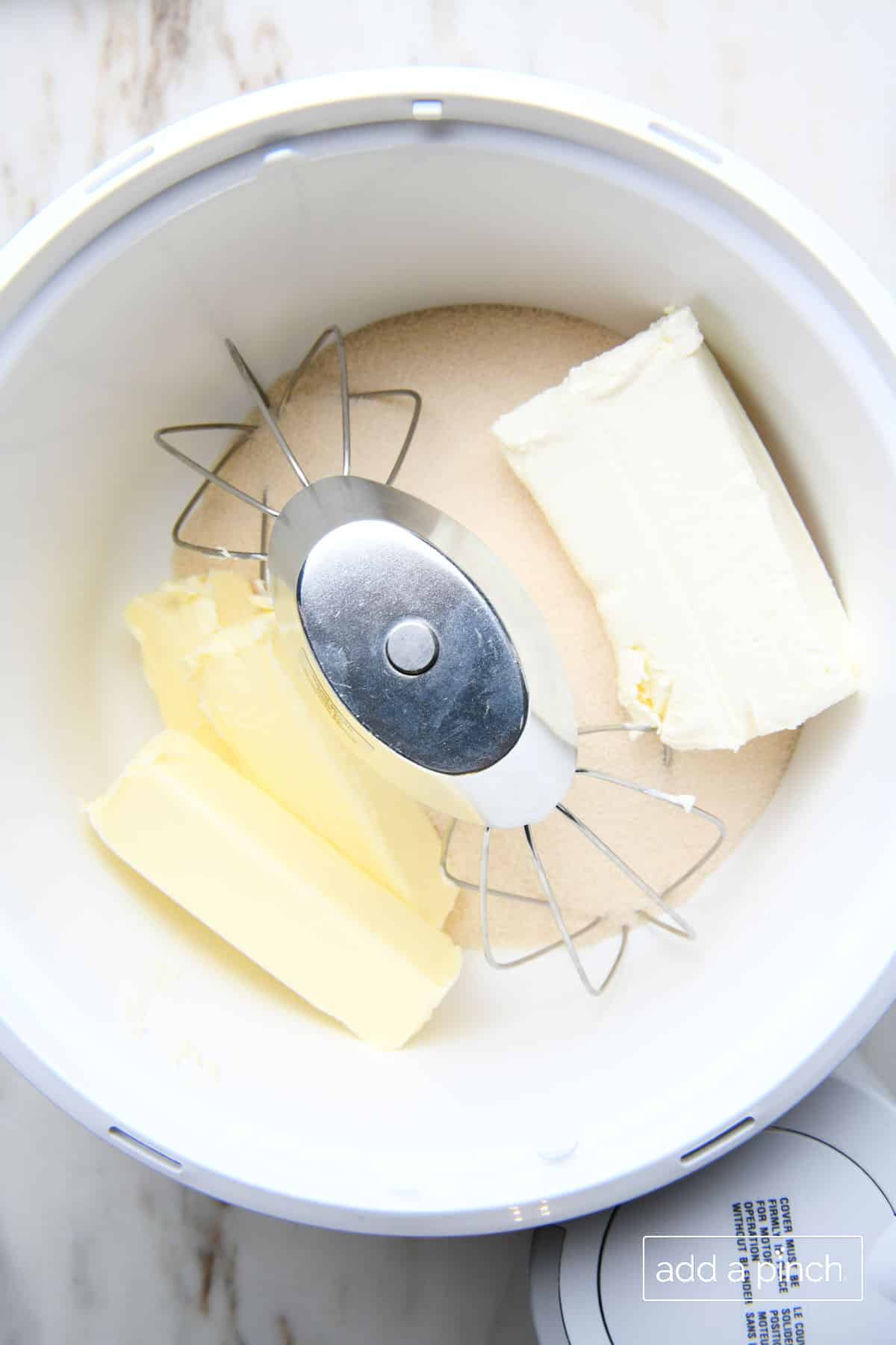 Stand mixer bowl with attached whisks holds sugar, block of cream cheese, and three sticks of butter. 