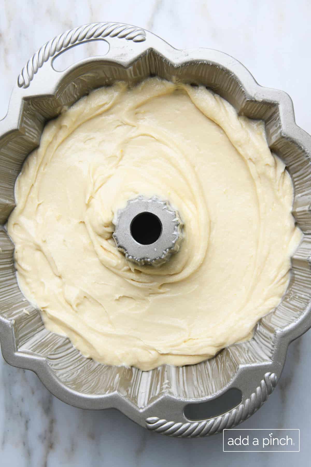 Lemon pound cake batter is poured into a greased and prepared silver bundt cake pan with handles. Pan sits on marble countertop.