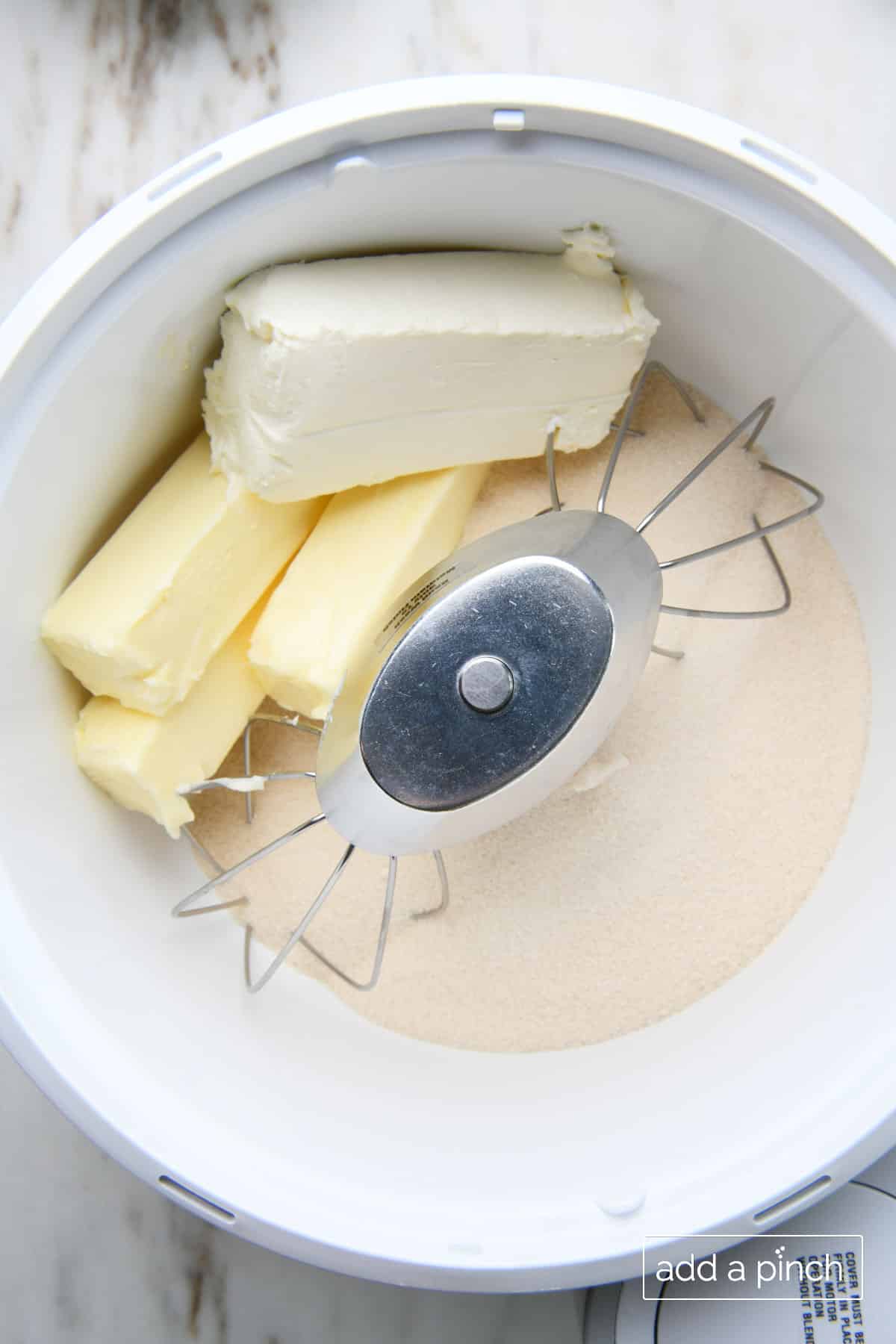 Bowl of a stand mixer holds block of cream cheese, three sticks of butter and sugar.