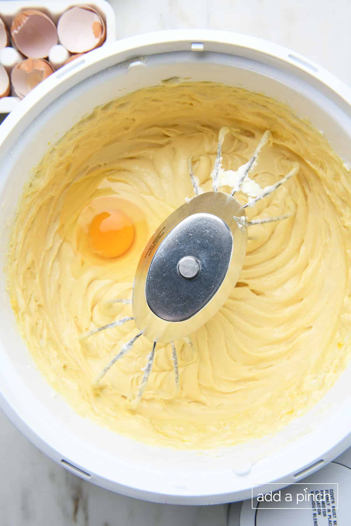 Final egg is blended into other mixed ingredients in a stand mixer bowl. Egg shells sit on marble countertop behind the bowl in egg container.