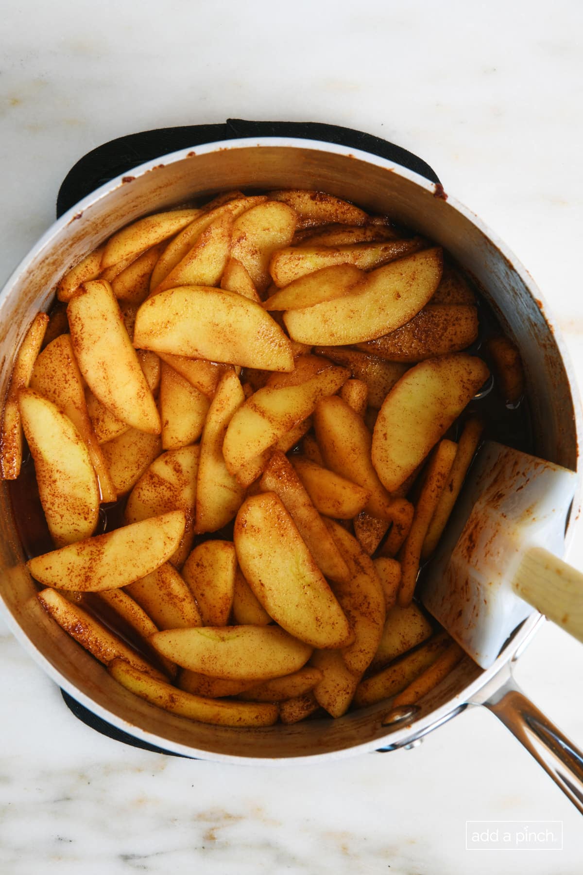 Apple slices are coated in cinnamon sauce mixture as it cooks in a saucepan and is stirred with a rubber spatula.