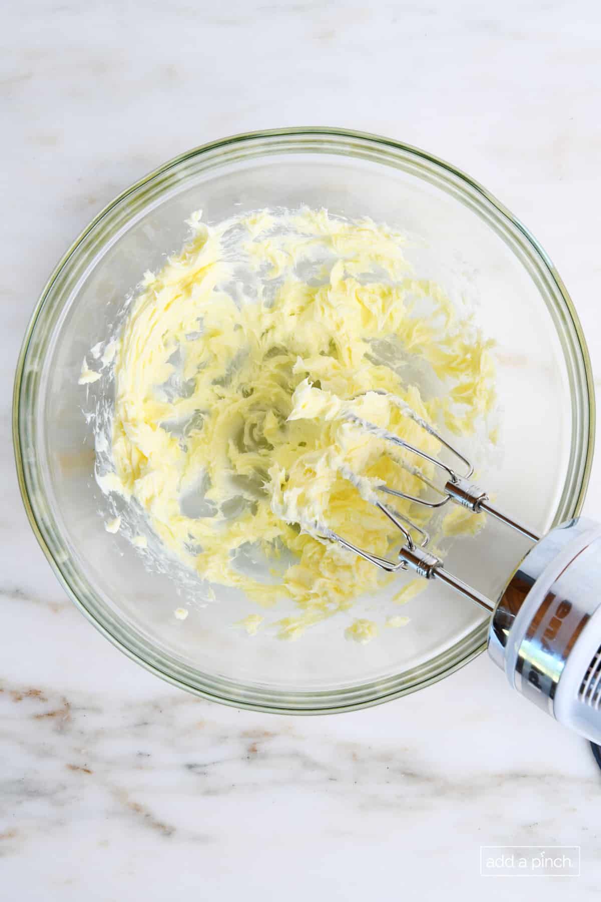 Softened butter is being creamed with a hand mixer in a glass bowl.