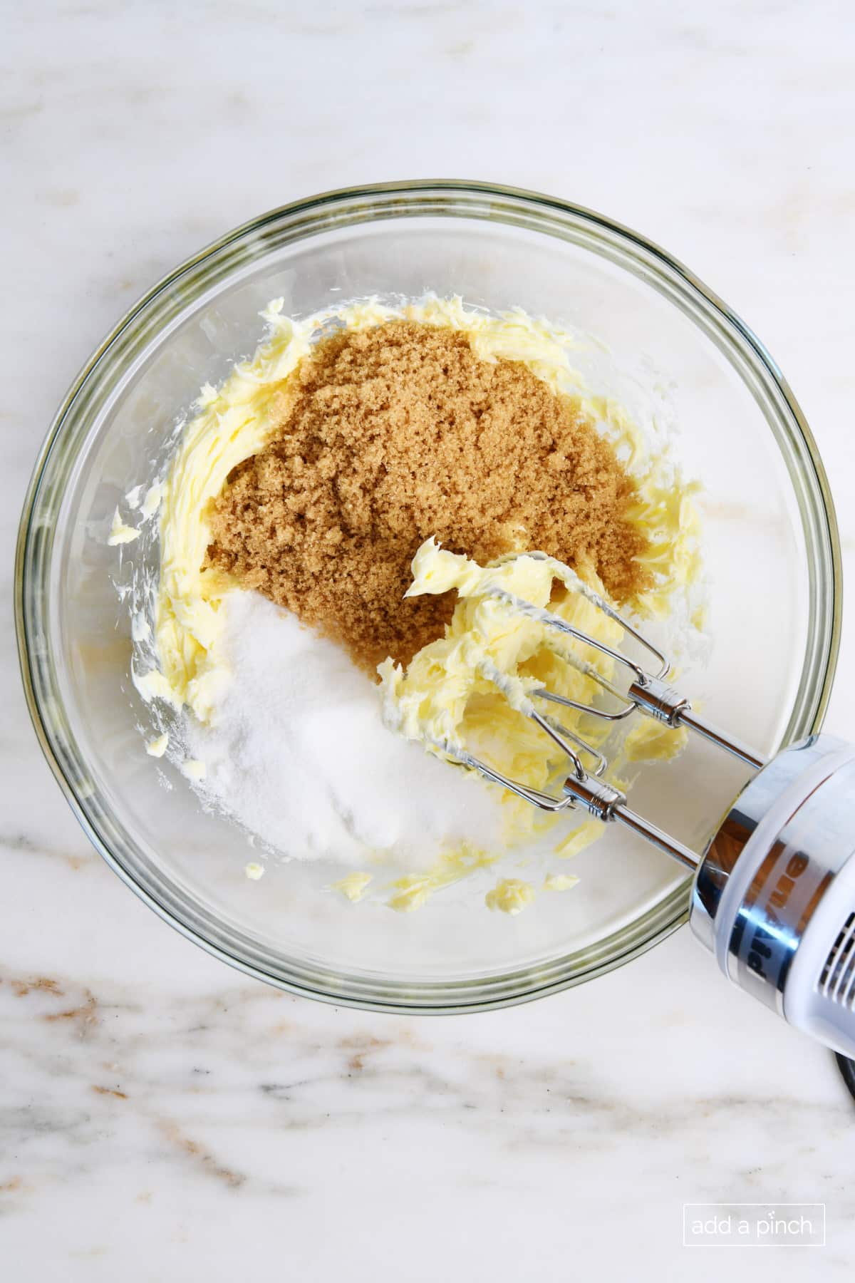 Glass mixing bowl holds softened butter, brown sugar and granulated sugar being creamed together with a hand mixer.