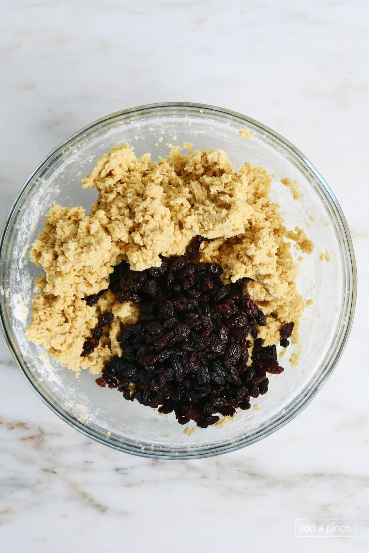 Glass bowl of cookie dough has raisins added ready to mix in to make oatmeal raisin cookie dough.