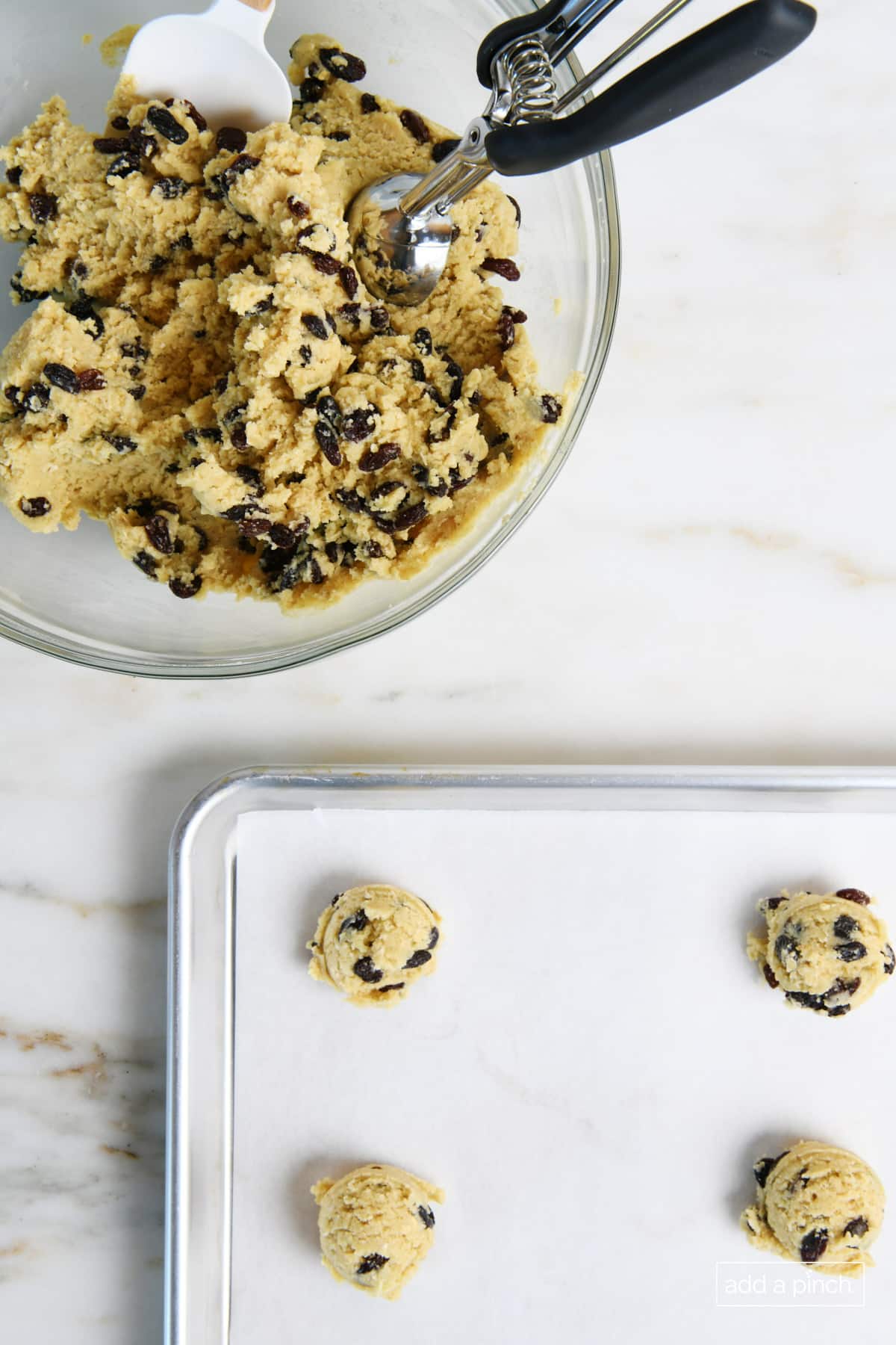 Oatmeal raisin cookie dough in glass bowl with scoop next to a parchment lined baking sheet with small scoops of cookie dough balls placed on it.