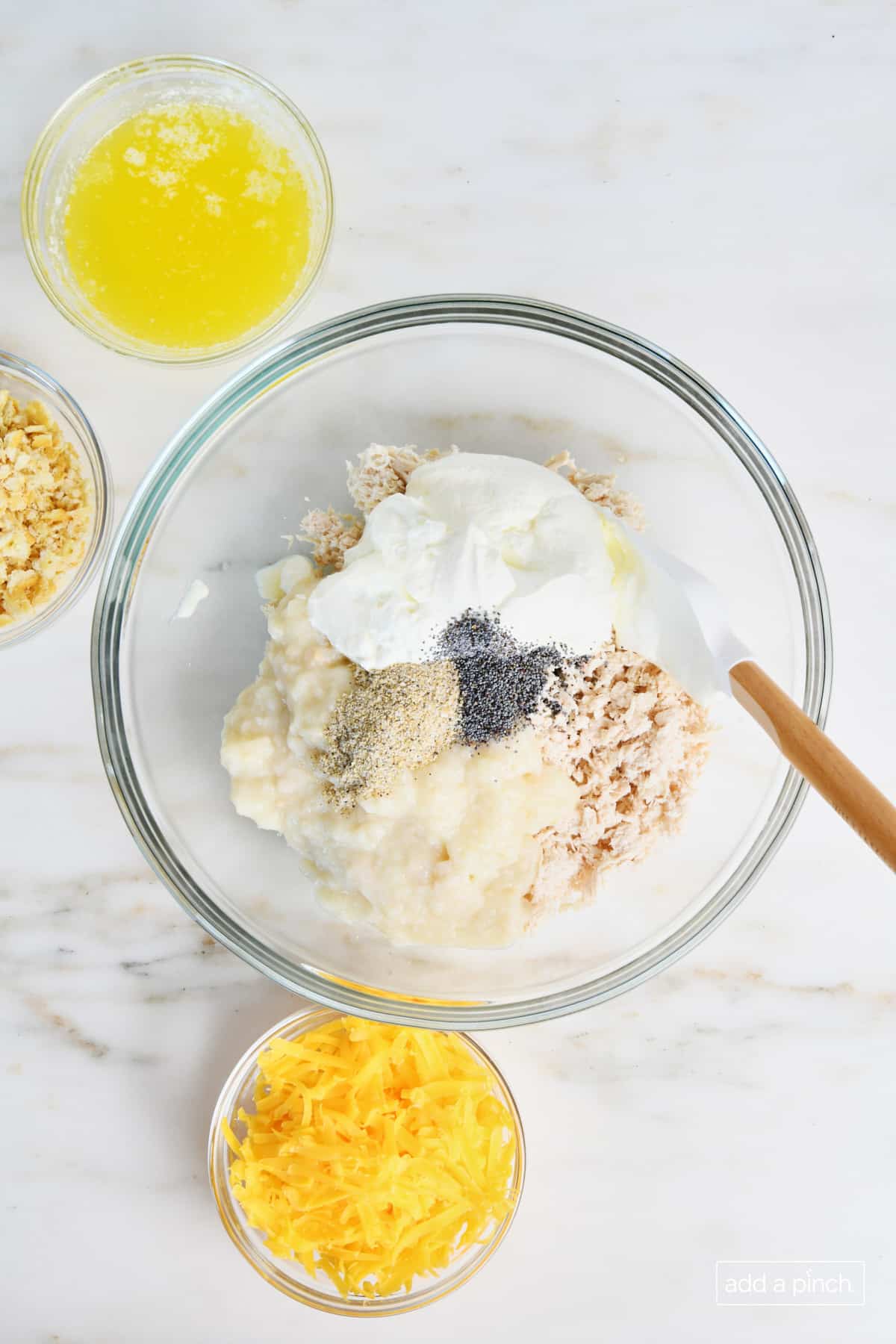 A glass bowl holds shredded chicken, sour cream, cream of chicken soup, poppy seeds and is surrounded by glass bowls of shredded cheddar, butter crackers and melted butter in preparation to mix together a chicken casserole. 