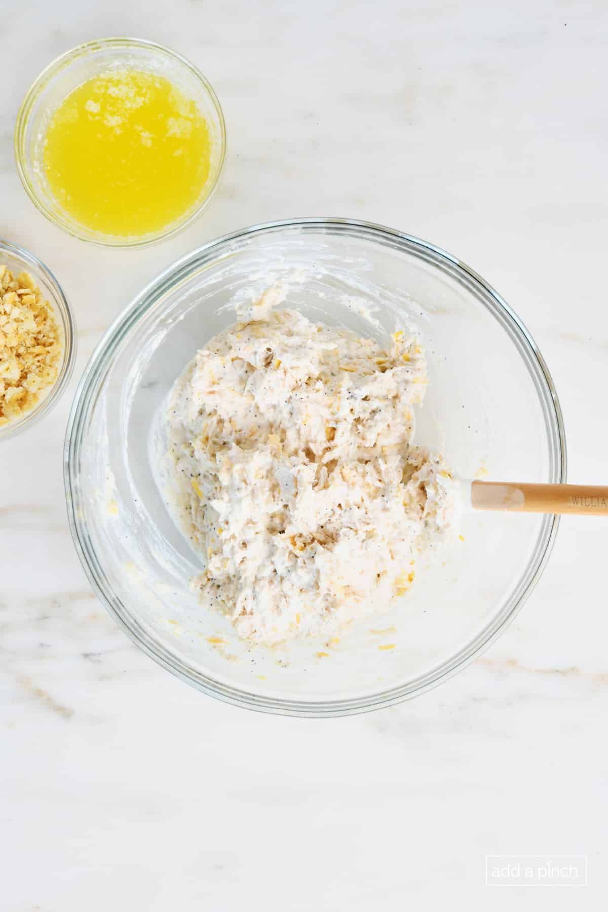 Glass mixing bowl with mixed ingredients for poppy seed chicken casserole. Surrounded by crushed butter crackers and melted butter on a white marble countertop. 