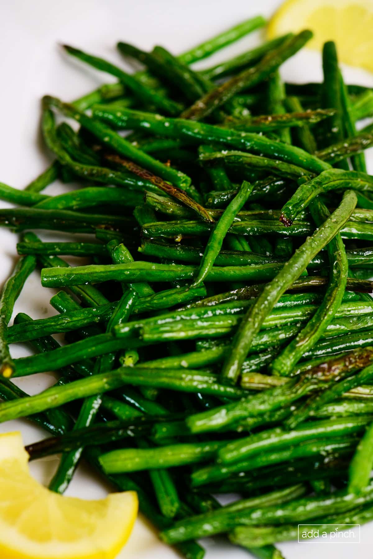 Closeup of roasted green beans showing caramelized areas of the crisp tender beans, flanked with lemon wedges on a white plate. 