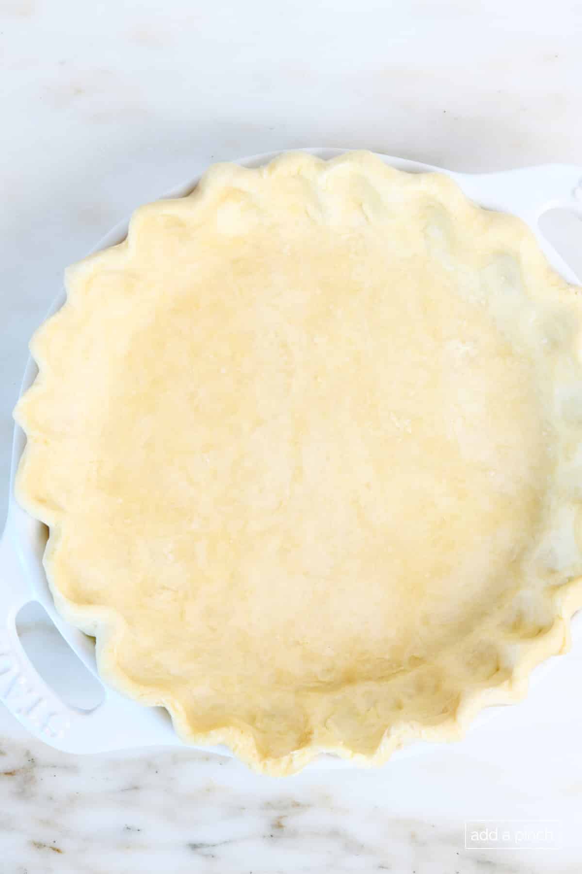 Pie crust has been baked to a flaky, golden color in a white pie dish on marble counterop.