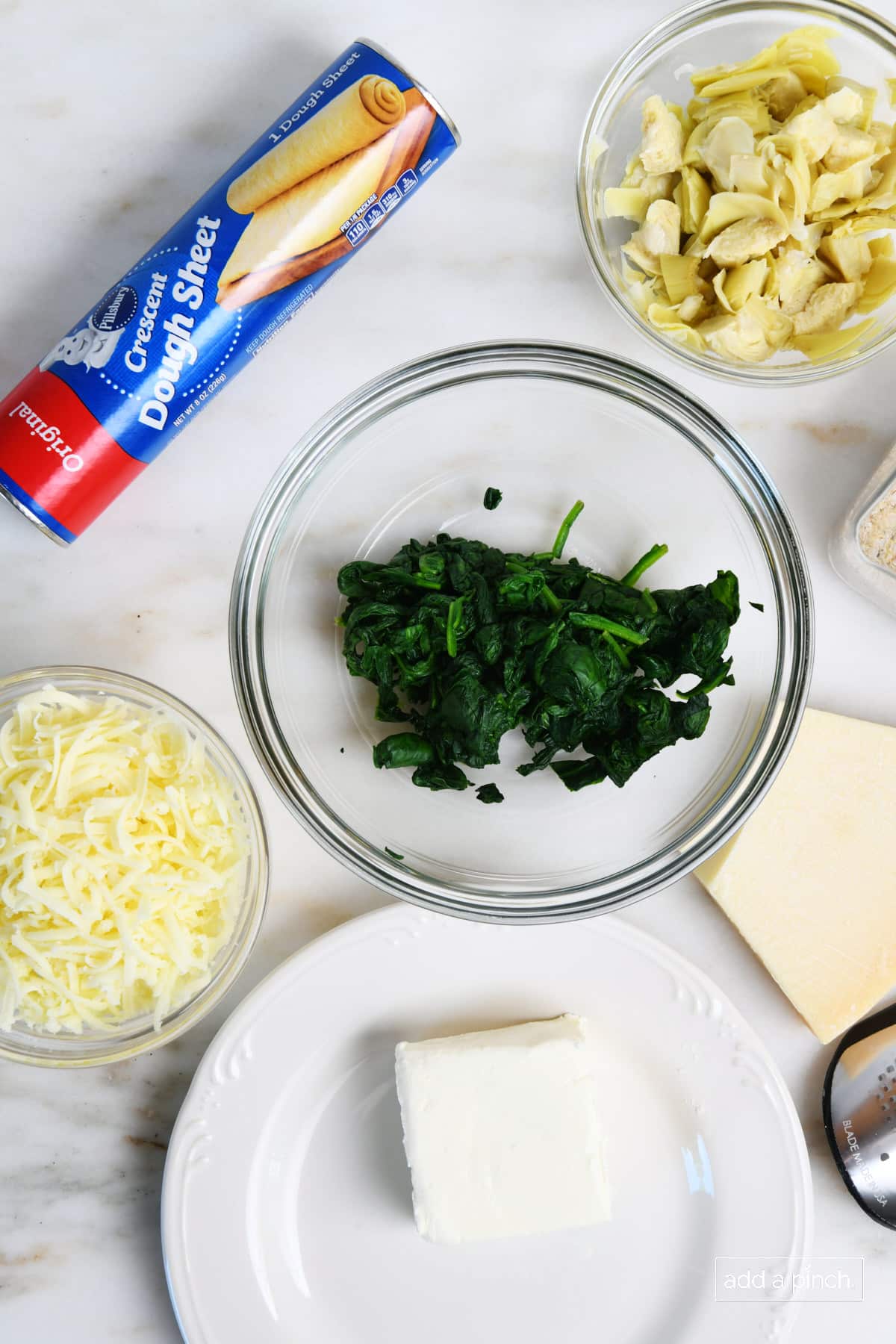 Spinach, artichokes, cheeses, a sheet of dough, and seasoning on a marble counter along with a cheese grater for making appetizers. 