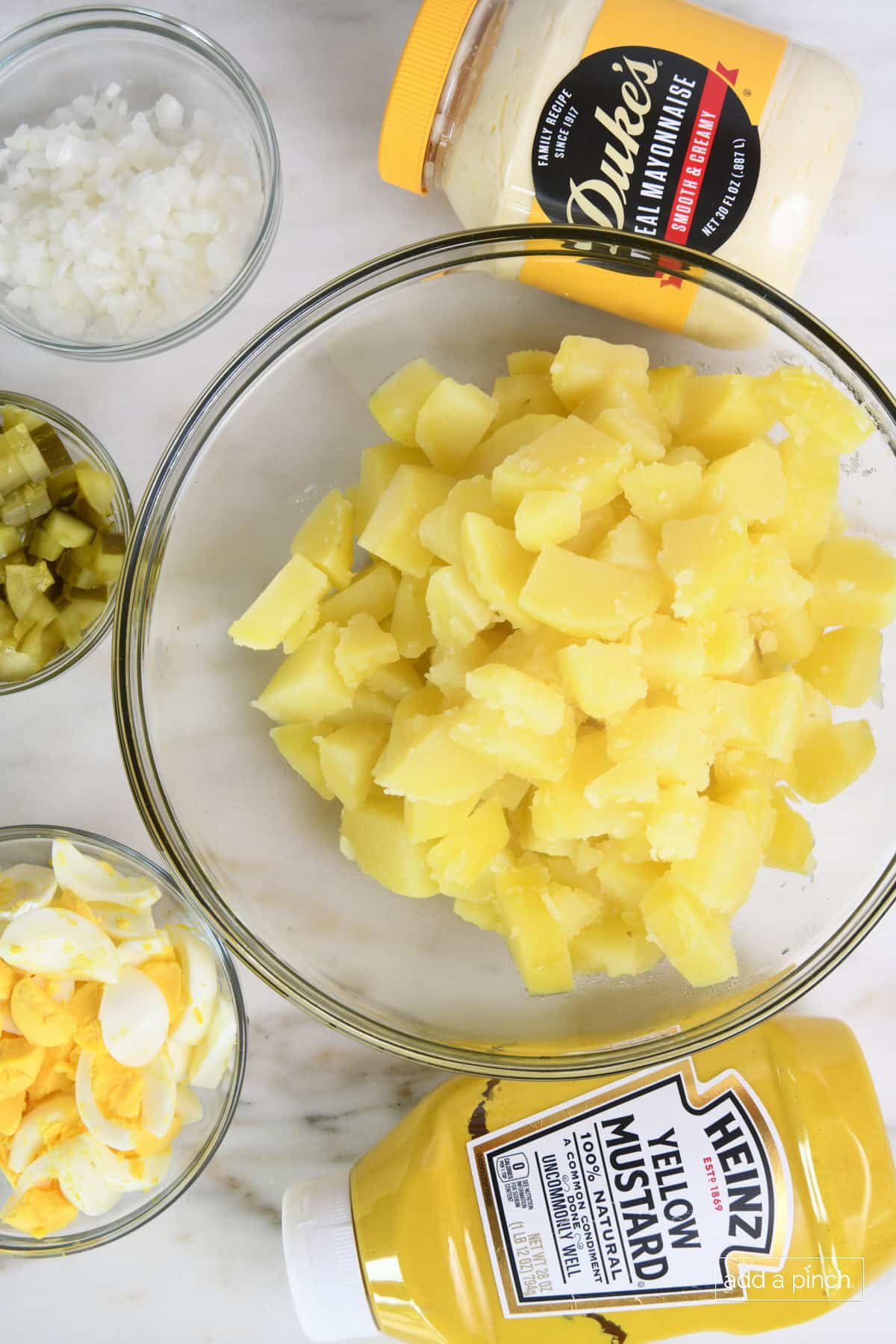 Glass bowls hold cooked cubed potatoes, diced onion, diced dill pickles, chopped eggs, mustard, mayo all on a countertop.