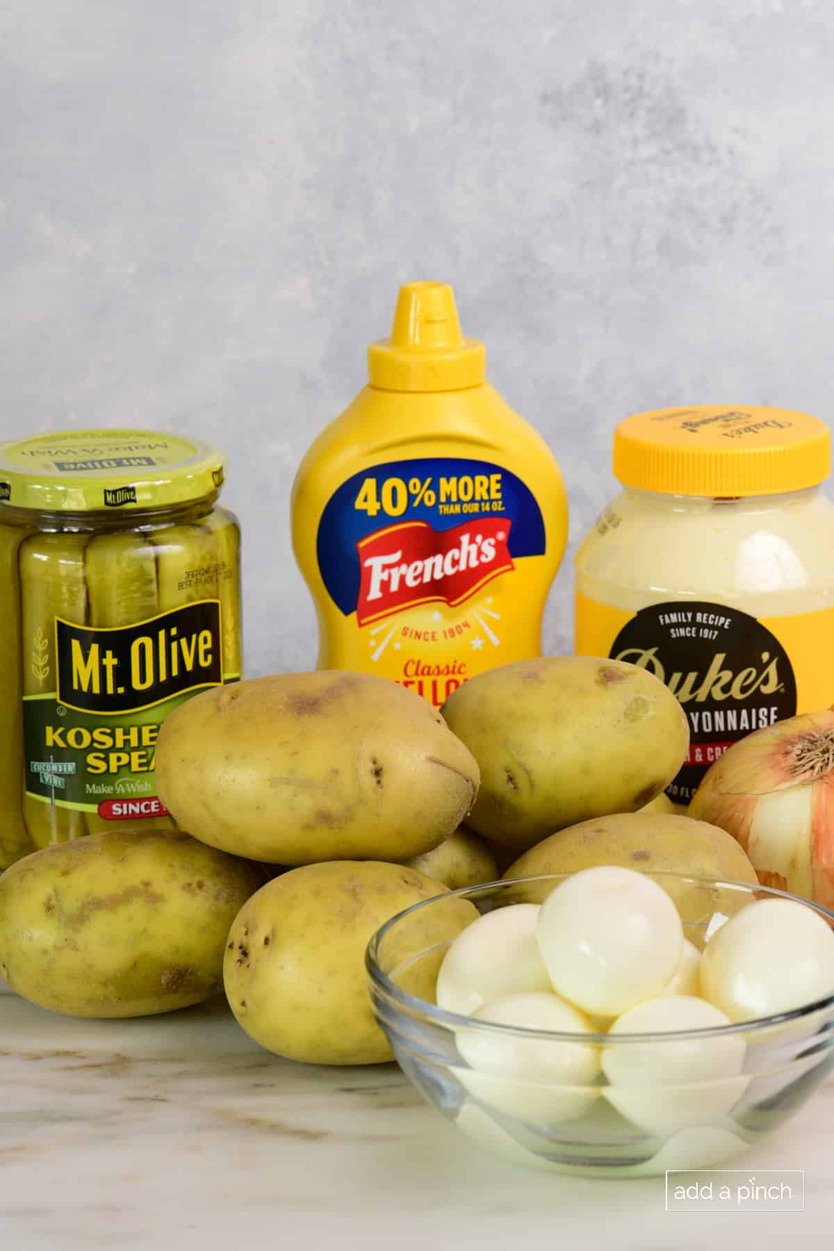 Russet potatoes, a jar of dill pickles, hardboiled eggs, an onion, mayo, and mustard sit ready to make this potato salad recipe.