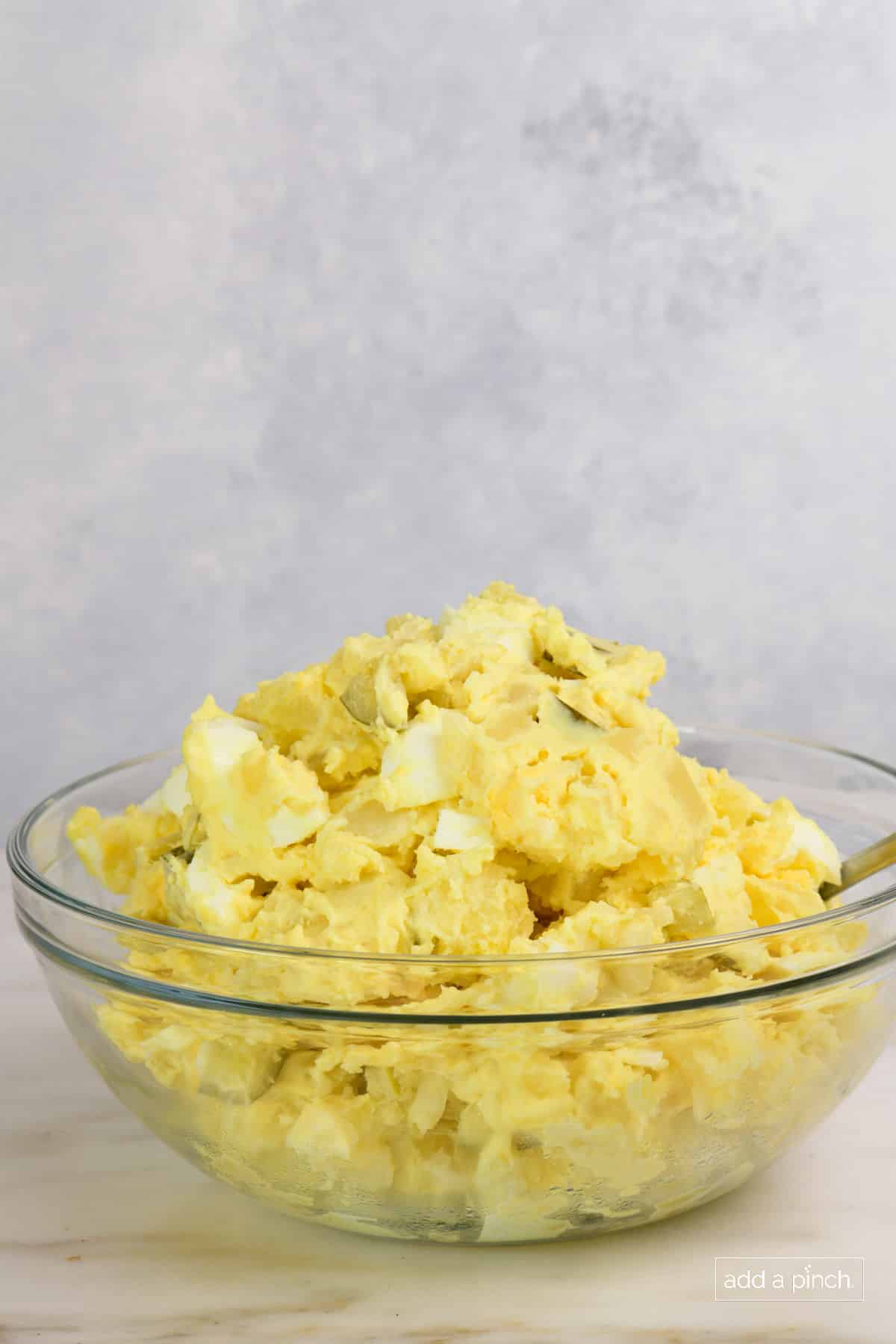 Side view of a big glass bowl full of potato salad that has eggs, mustard, pickles visible. 