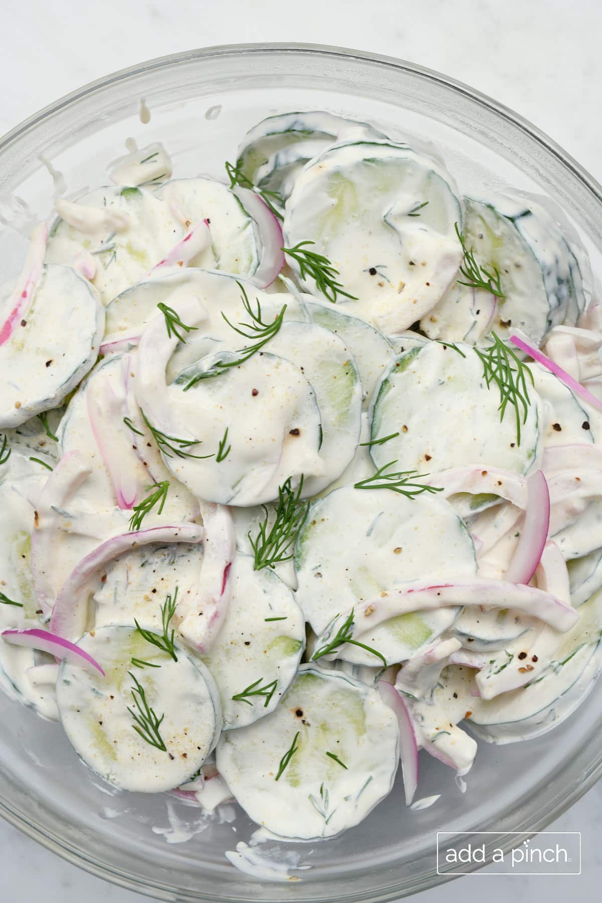 Sliced cucumbers, red onion, dill and a creamy dressing in a glass bowl.