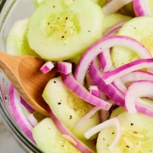 Glass bowl with cucumbers and red onion in cucumber salad with a wooden spoon.