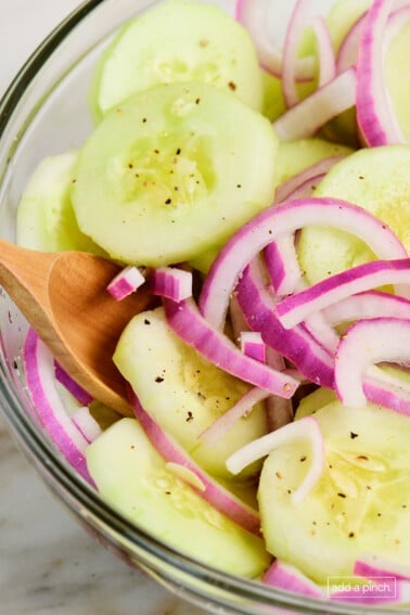Glass bowl with cucumbers and red onion in cucumber salad with a wooden spoon.