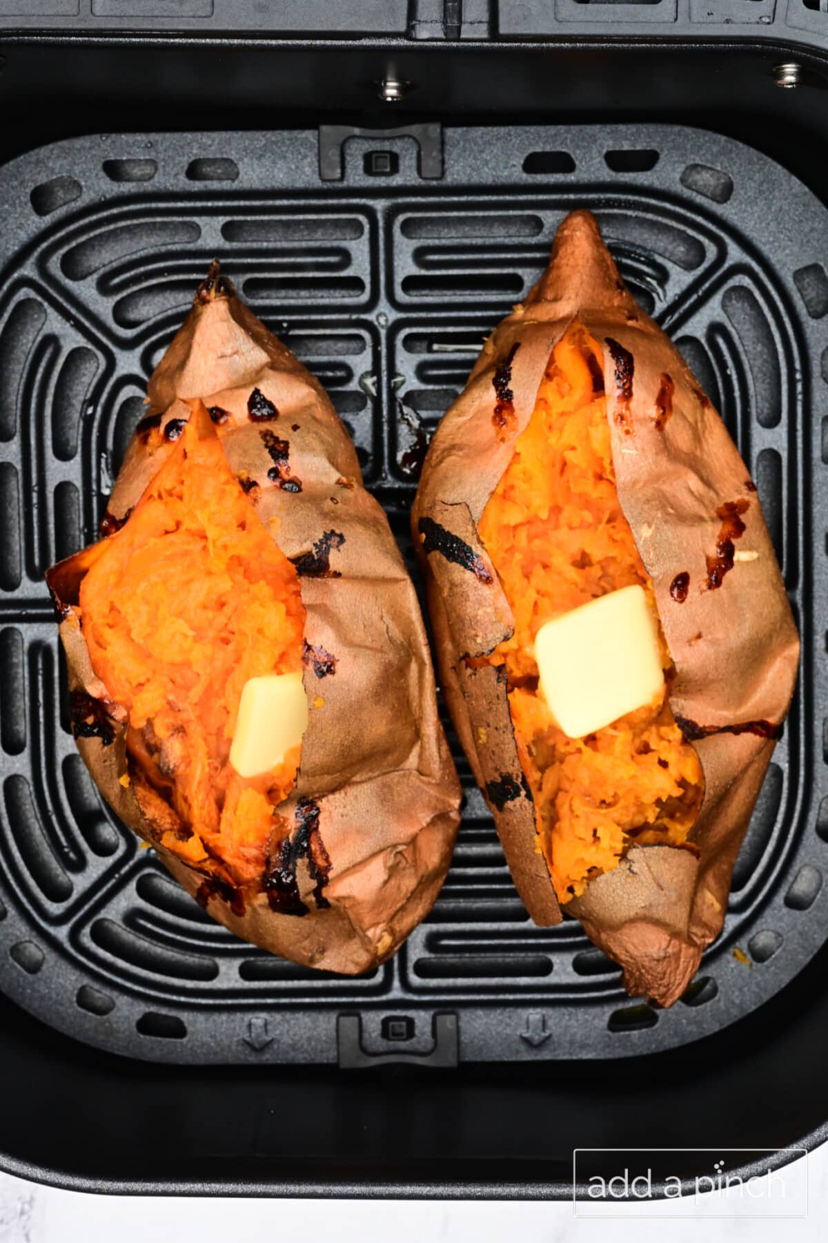 Two air fryer baked sweet potatoes in an air fryer basket. Both are opened to show the fluffy inside with a pat of butter.