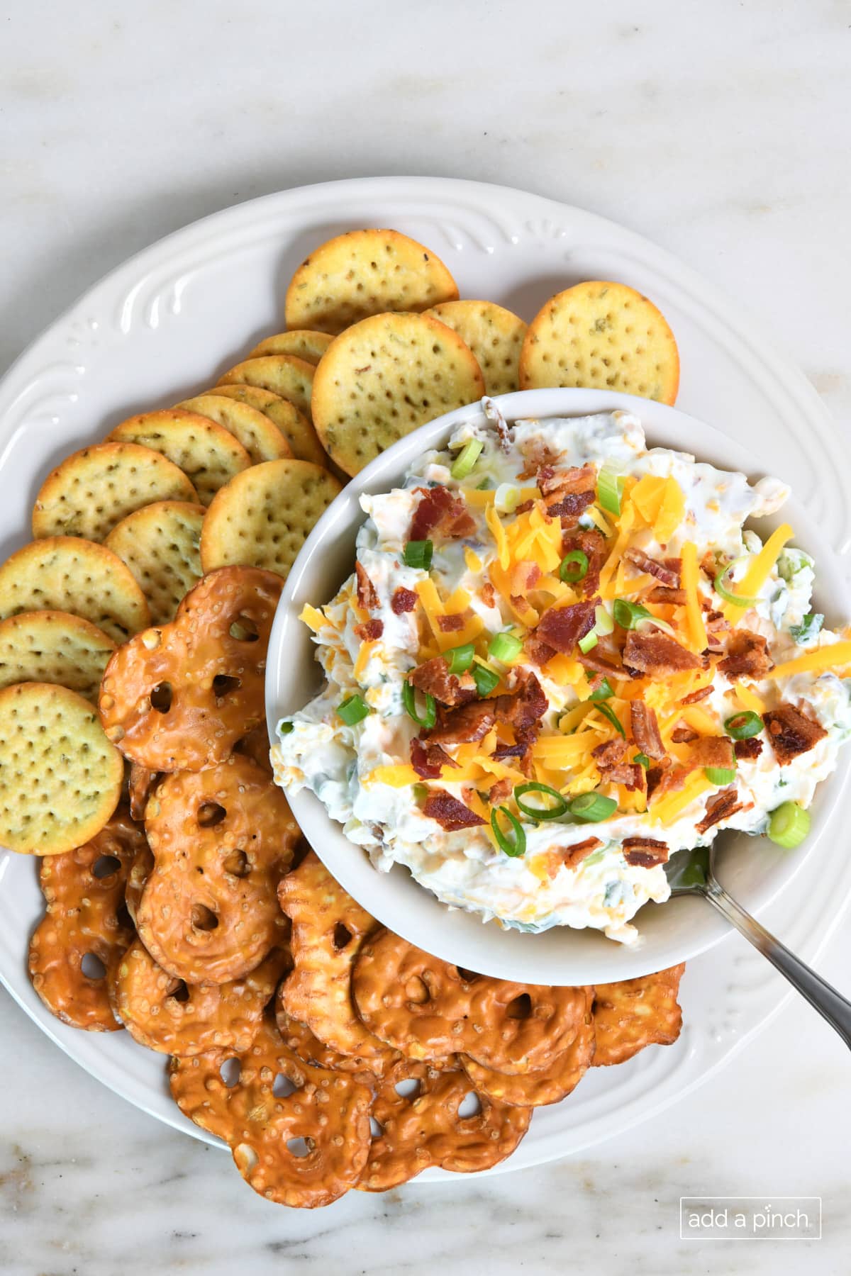 Bacon cheddar dip topped with cheese, bacon, and green onions on a platter with crackers and pretzel chips.