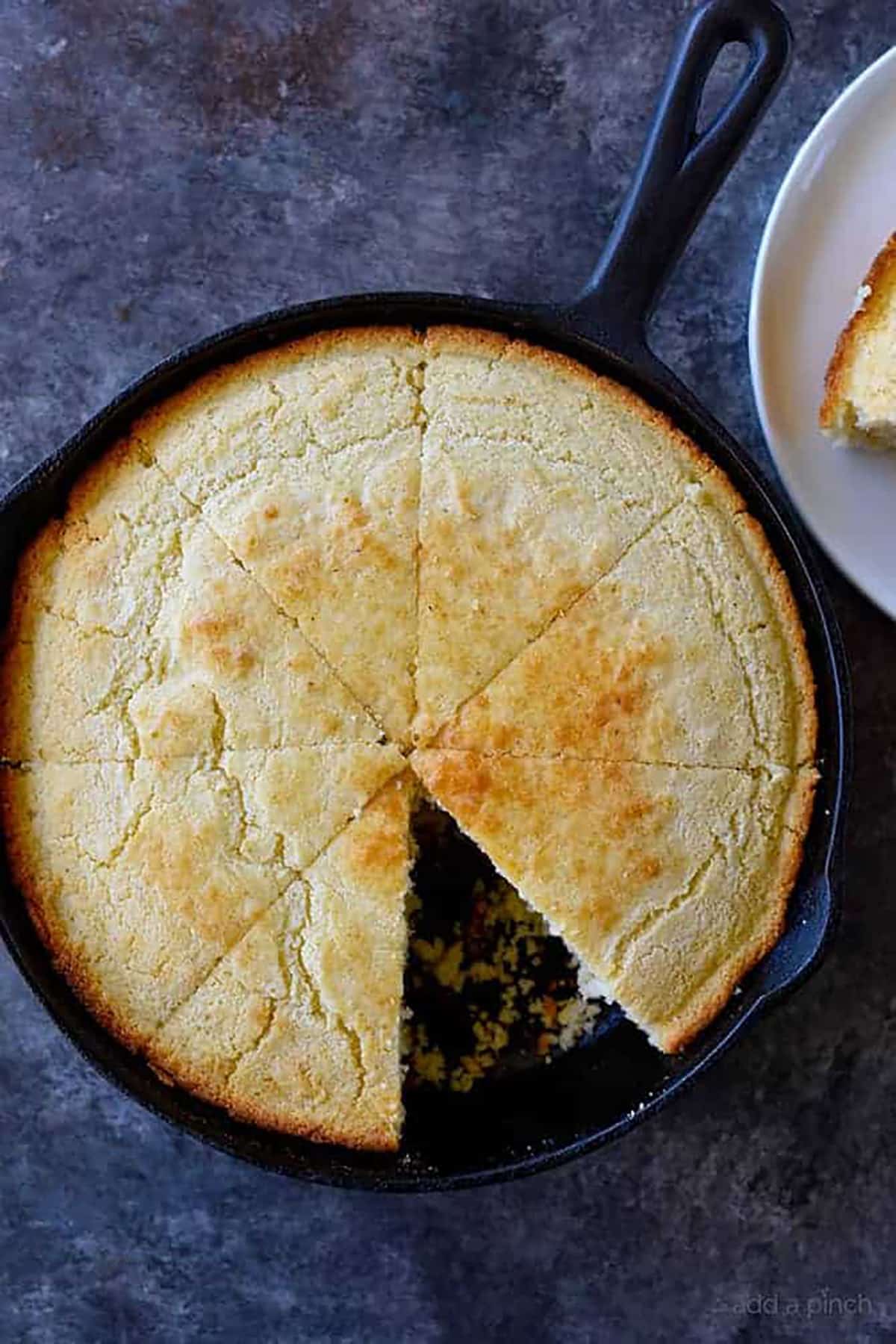 Cast iron skillet holds golden brown southern cornbread sliced into wedges with one wedge served on a plate.