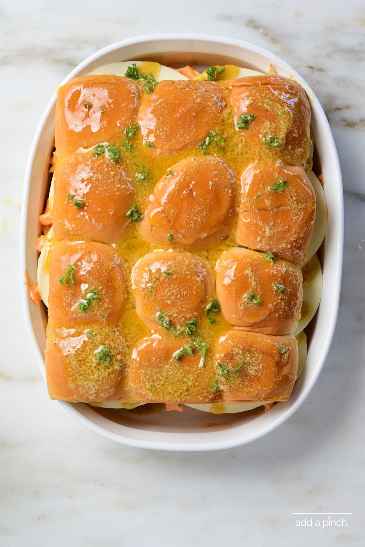 Buffalo chicken sliders topped with buttery spread in a white baking dish.