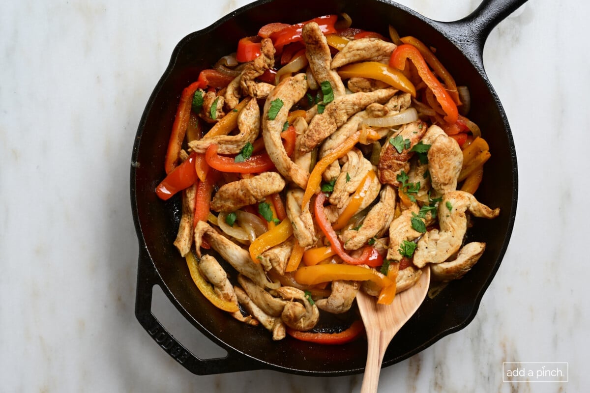 Chicken, pepper strips and other ingredients for Chicken Fajitas in a cast iron skillet with a wooden spoon.