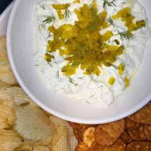 Dill pickle dip served with ruffled potato chips, pretzels, and fresh veggie sticks on a sunny outdoor table perfect for summer lake days or game day.