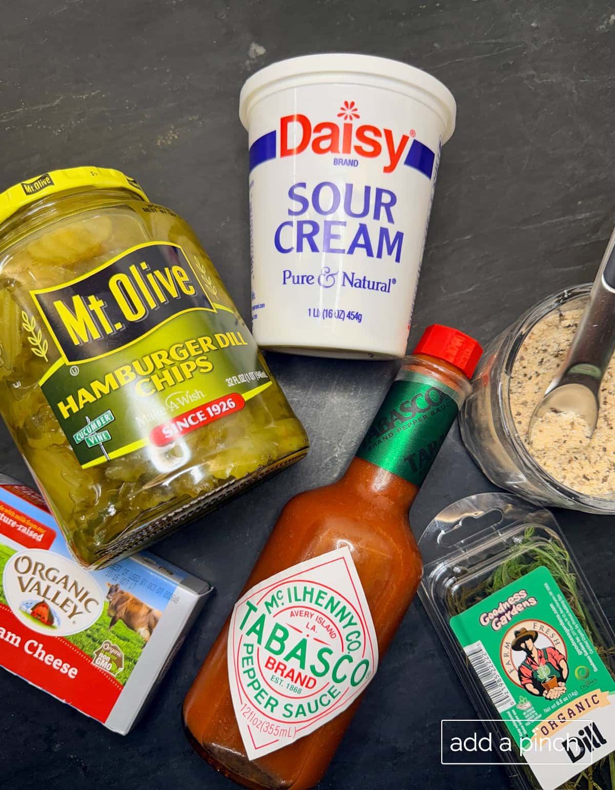 Flat lay of ingredients for the best dill pickle dip recipe: cream cheese, sour cream, chopped dill pickle chips, fresh dill, Stone House Seasoning, and hot sauce on a wooden kitchen counter.